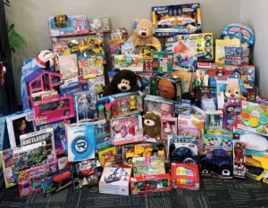 A large pile of assorted new toys for all ages, including dolls, games like Monopoly and Battleship, building sets, stuffed animals, and balls, gathered for a donation or toy drive.