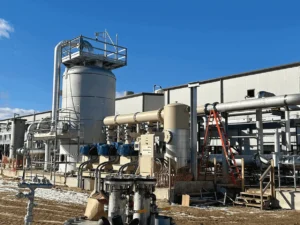 An industrial gas or processing plant featuring large, light-colored vertical tanks, horizontal pipes, and a metal building under a bright blue sky. An orange ladder leans against one of the pipes.