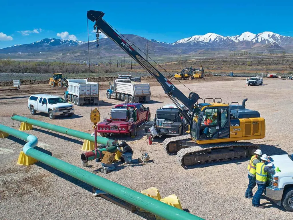 Pipeline fabrication underway on a gravel laydown yard, showing workers welding pipe sections next to a heavy excavator and several dump and support trucks, with mountains in the distance.