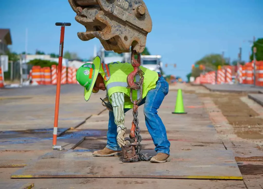 Close-up of a worker in personal protective equipment (PPE) bending over on a construction site to rig up a load for a crane or excavator, with temporary safety barriers lining the road in the background.