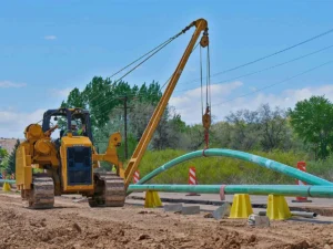 A large, yellow pipelayer side boom tractor uses its crane arm to carefully bend a section of coated green steel pipeline over support stands on a dirt road construction site.
