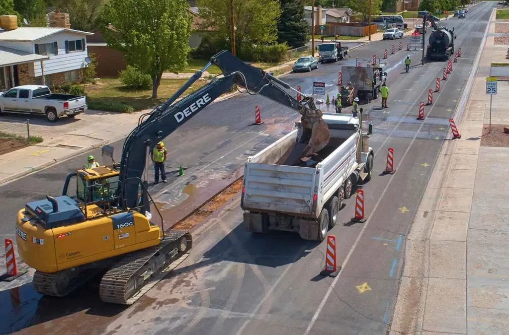 Road construction and paving preparation on a residential street, showing an excavator removing road material, with traffic cones, other equipment, and a vacuum truck lined up down the street.