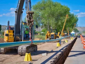 A large excavator operator uses the machine to lift and position a light-blue coated pipeline section over an open trench, with other side boom equipment and workers lined up in the background.