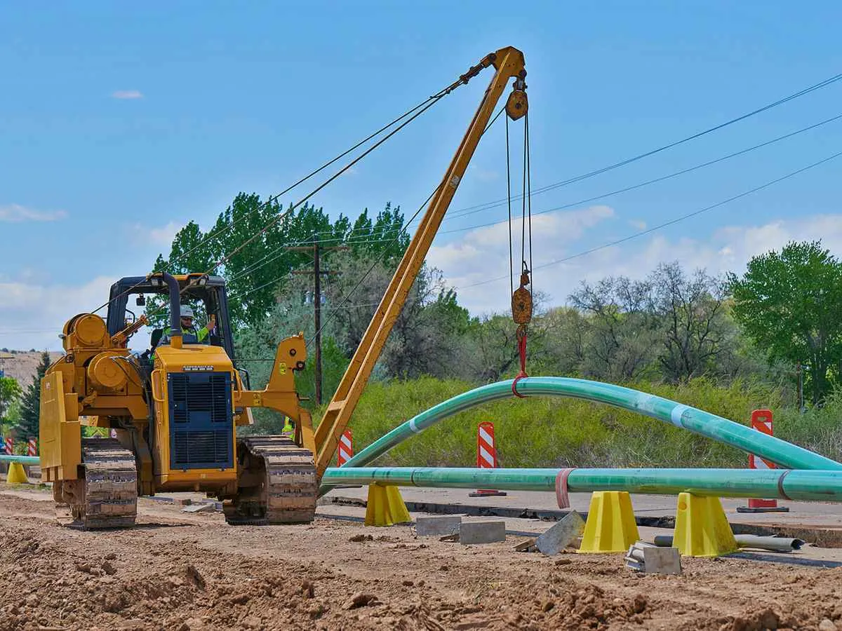 A large, yellow pipelayer side boom tractor uses its crane arm to carefully bend a section of coated green steel pipeline over support stands on a dirt road construction site.