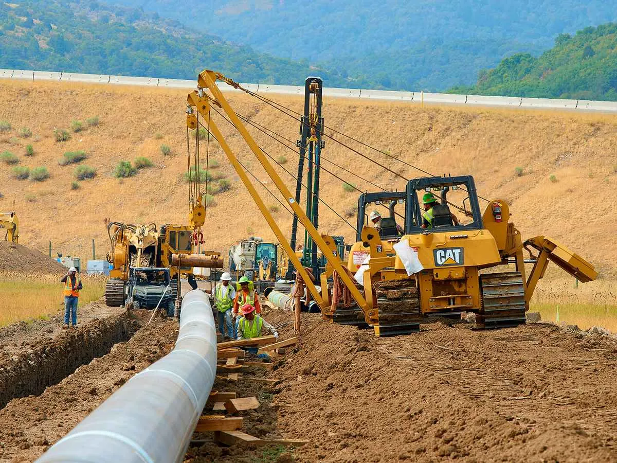 Heavy equipment, including a yellow CAT pipelayer tractor and an excavator, performing the lowering-in phase of a pipeline installation project near a hillside.