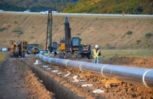 A construction crew preparing to install a large-diameter steel pipeline in an open trench, with heavy machinery and workers visible.