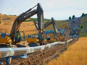 A large-diameter oil or gas pipeline being installed across a dry, grassy hillside. Several heavy excavators (Canyon Pipeline 350G) are positioned to lower the pipe section, while construction workers assist along the trench line.