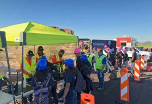 A group of students wearing blue and pink hard hats are gathered under a lime green canopy listening to construction workers in high-visibility gear at an outdoor educational or career event in a desert setting.