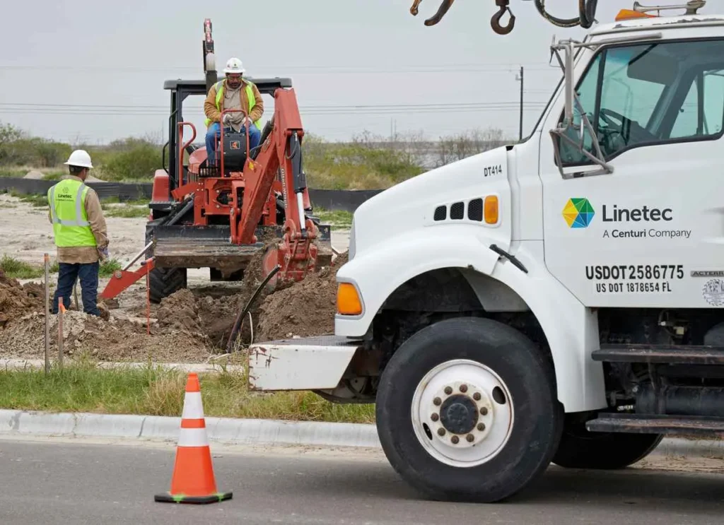 A close-up of the front of a white utility truck, labeled 'Linetec,' parked near a roadside. In the background, two construction workers in safety gear oversee a small, red backhoe digging a trench in the dirt.
