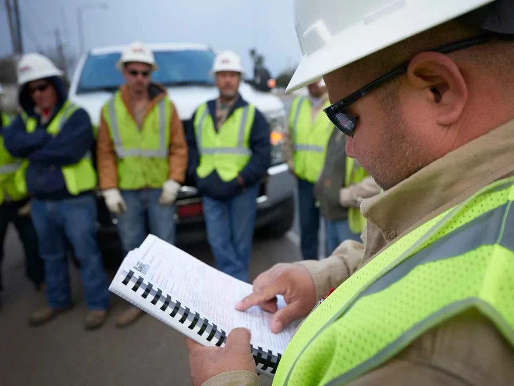 A close-up, over-the-shoulder shot of a construction supervisor in a hard hat and safety vest reading and pointing at a safety document or plan book. A crew of five other workers in safety gear stands blurred in the background, facing the supervisor.
