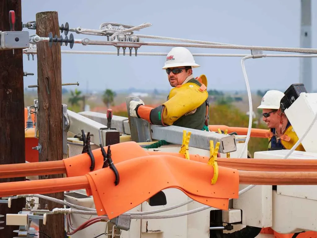 Two utility linemen in protective gear, including hard hats, safety glasses, and yellow insulating sleeves, working from a bucket truck high near power lines and a utility pole. The worker in the foreground is looking left.