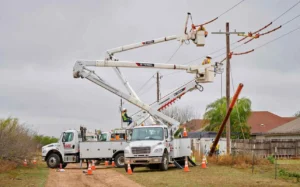 Two utility workers are elevated in the buckets of two large white boom trucks, performing maintenance on power lines near a utility pole and residential area. A broken utility pole lies angled to the right.
