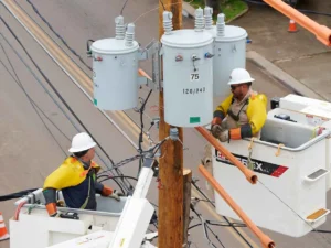 Two utility linemen in hard hats and yellow protective sleeves are elevated in bucket trucks, working on a cluster of three large electrical transformers mounted on a wooden utility pole, with many power lines surrounding them.
