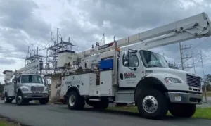 Two large white utility bucket trucks, both labeled 'Linetec Services,' are parked on an asphalt road with their booms extended, facing a high-voltage electrical substation under a cloudy sky.