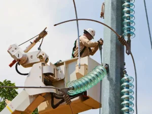 A utility worker in a hard hat and harness is elevated in a bucket truck, working on a large metal utility tower. The scene is dominated by a stack of blue-green glass insulators and a bent metal jumper wire near the worker.