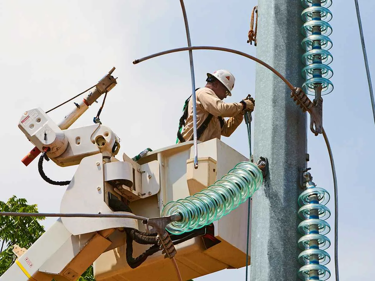 A utility worker in a hard hat and harness is elevated in a bucket truck, working on a large metal utility tower. The scene is dominated by a stack of blue-green glass insulators and a bent metal jumper wire near the worker.