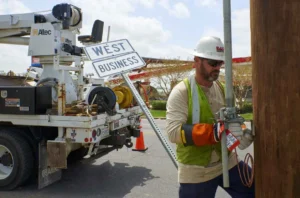 A utility worker in a hard hat, safety vest, and protective gloves is attaching a metal ground rod to a wooden utility pole. A white Altec boom truck is parked behind him, carrying a 'WEST BUSINESS' road sign.
