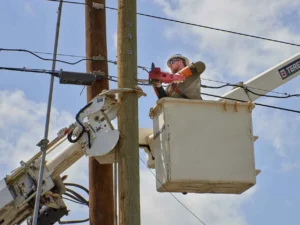A utility worker in a hard hat, safety glasses, and protective sleeves is elevated in the white bucket of a boom truck, using a specialized hydraulic tool to work on equipment attached to a wooden utility pole.