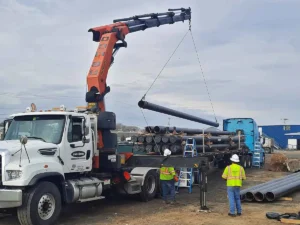 A crane truck with an orange articulating boom is lifting a large black pipe off a flatbed trailer stacked with many similar pipes. Two construction workers in hard hats and high-visibility vests supervise the pipe rigging and lifting operation.