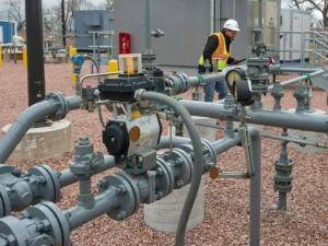 A technician or engineer in an orange safety jacket, a black hard hat, and safety glasses is operating controls on a large grey industrial electrical panel indoors.