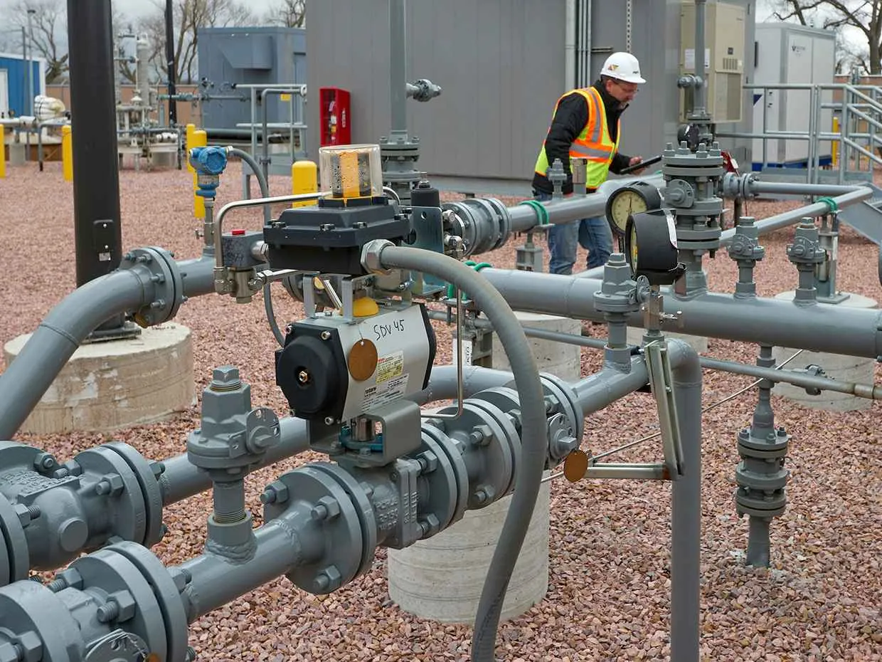 A technician or engineer in an orange safety jacket, a black hard hat, and safety glasses is operating controls on a large grey industrial electrical panel indoors.