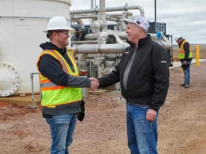Two men, one wearing a white hard hat and a high-visibility vest and the other in a black jacket and baseball cap, shake hands and smile in front of a natural gas or biogas processing facility with large metal tanks and complex piping.