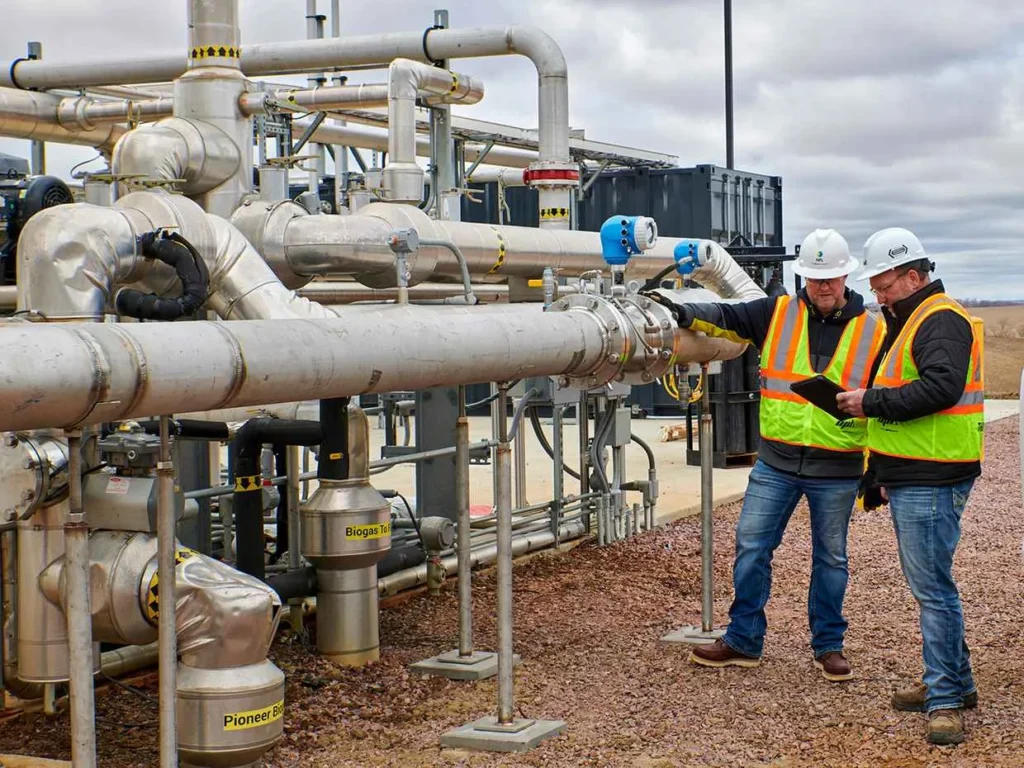 Two engineers or workers in white hard hats and high-visibility vests are inspecting a complex network of silver industrial pipes and valves outdoors at a processing plant, with one holding a tablet.