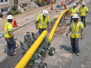 Utility workers in high-visibility vests and hard hats monitor a hydraulic machine fusing two sections of large, yellow polyethylene (PE) gas pipe on a residential sidewalk and street. Orange traffic cones and warning tape are visible in the background.