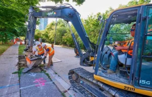 Three construction workers are operating an excavator to dig a trench along a city sidewalk. One worker is driving the yellow machine, and two others in hard hats and high-visibility vests are guiding the excavation bucket.