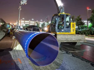 A large, dark blue pipe with a white band is lowered into a trench at night by a yellow and gray John Deere excavator. The pipe opening is visible in the foreground, and the background shows a paved roadway and a brightly lit parking garage.