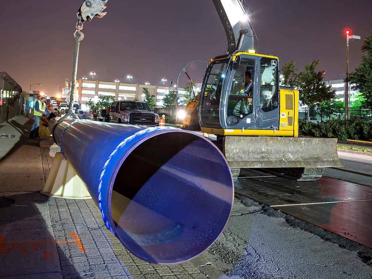 A large, dark blue pipe with a white band is lowered into a trench at night by a yellow and gray John Deere excavator. The pipe opening is visible in the foreground, and the background shows a paved roadway and a brightly lit parking garage.