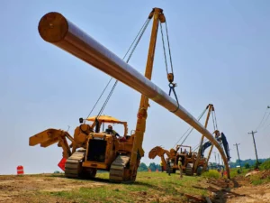 Two Caterpillar pipelayers, or sidebooms, operate in tandem to lift and position a long, large-diameter section of steel pipe for pipeline construction on a sunny day.