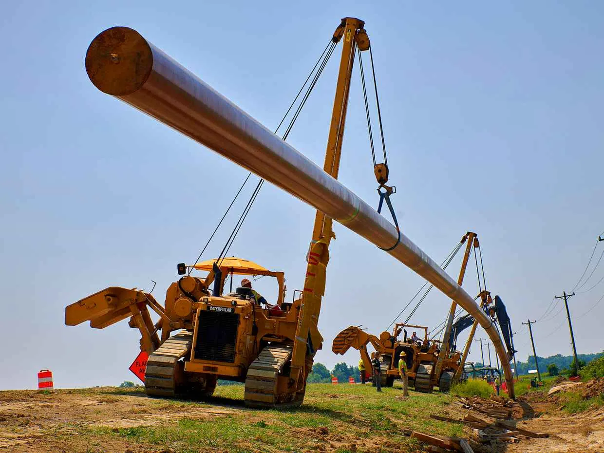 Two Caterpillar pipelayers, or sidebooms, operate in tandem to lift and position a long, large-diameter section of steel pipe for pipeline construction on a sunny day.