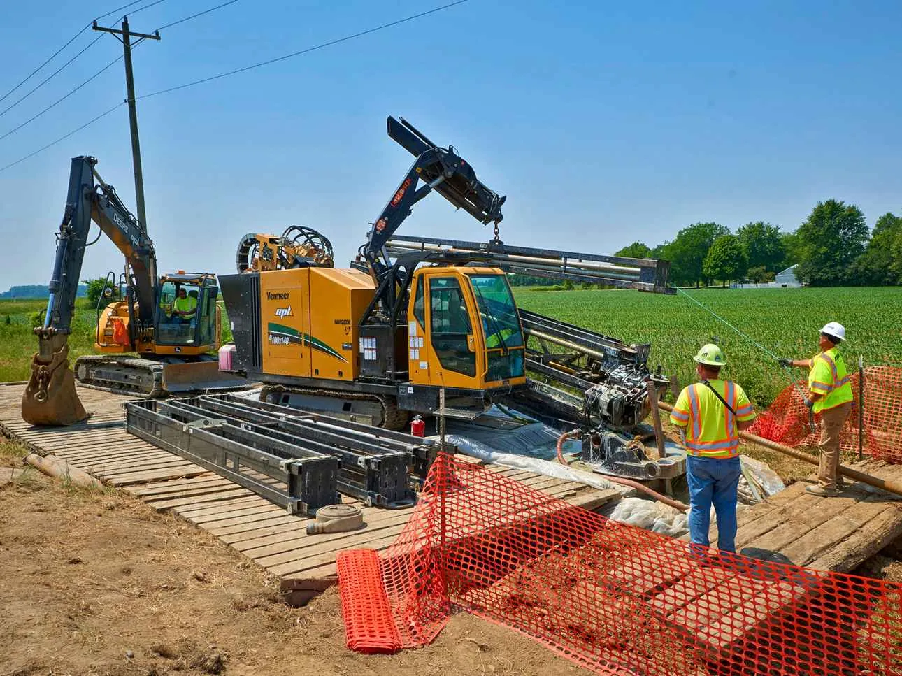 A large, yellow Vermeer D220x300 horizontal directional drill (HDD) and a John Deere excavator operate on wooden construction mats in a rural field under a bright sun. Two workers in hard hats and safety vests stand in the foreground, supervising the drilling operation near orange safety fencing.
