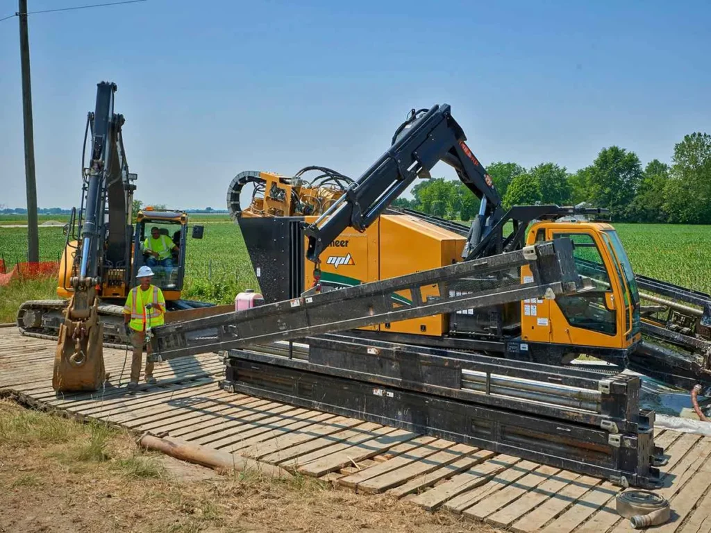 An outdoor utility construction scene showing a yellow Vermeer horizontal directional drilling machine and a smaller excavator resting on a temporary wooden access mat. A worker in a white hard hat and safety vest stands in the foreground.
