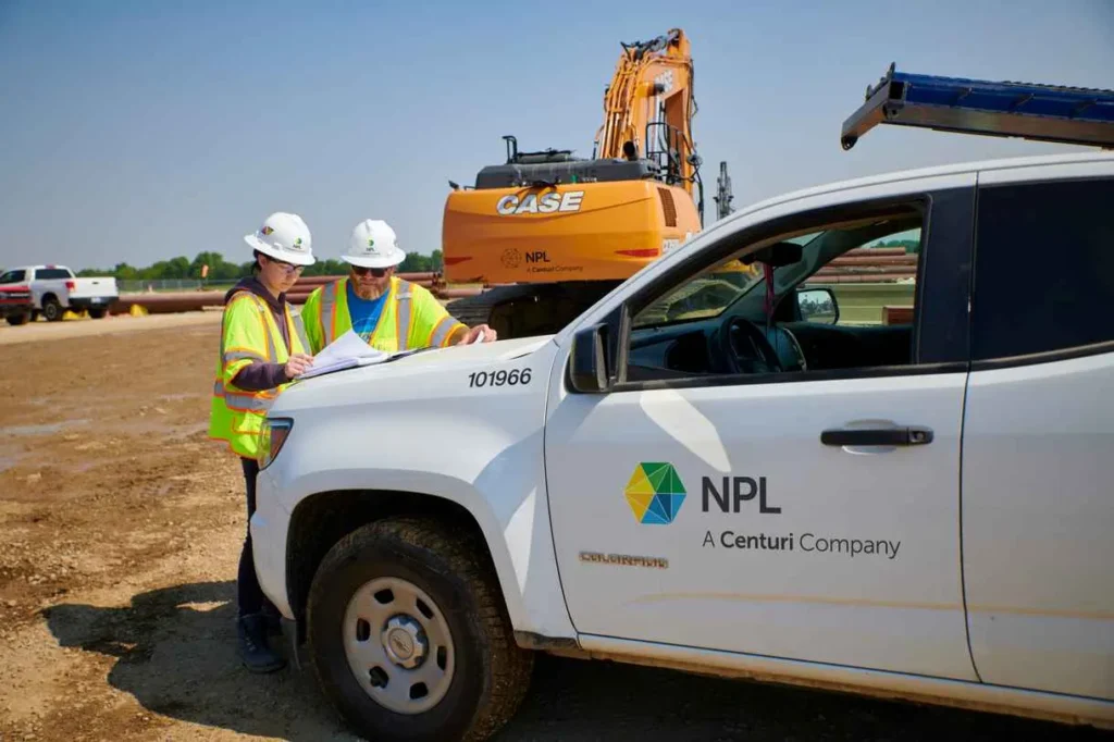 Two construction workers in safety vests and hard hats look at a large blueprint or map resting on the hood of a white NPL A Centuri Company pickup truck at a construction site. An orange CASE excavator is visible in the dirt background.
