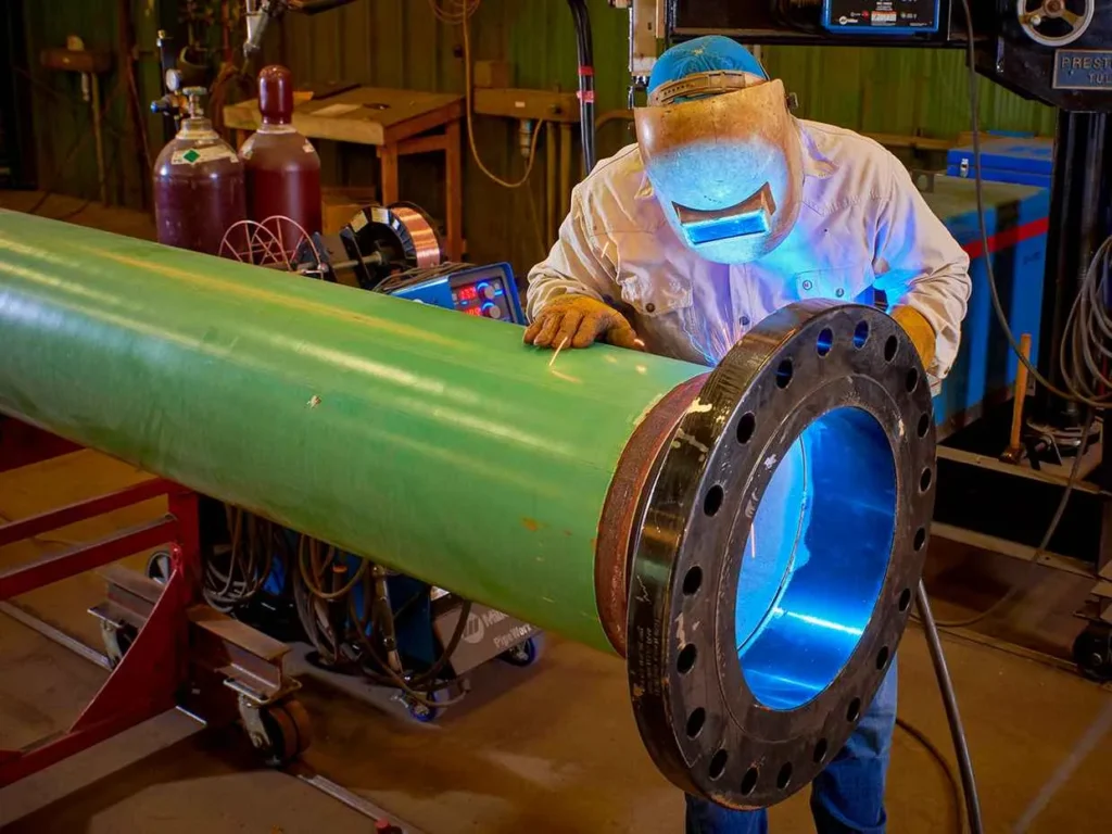 A worker wearing a welding helmet and gloves welds a large, heavy steel flange onto the end of a long, bright green coated pipe inside a fabrication shop. The bright blue arc of the welding process is visible.