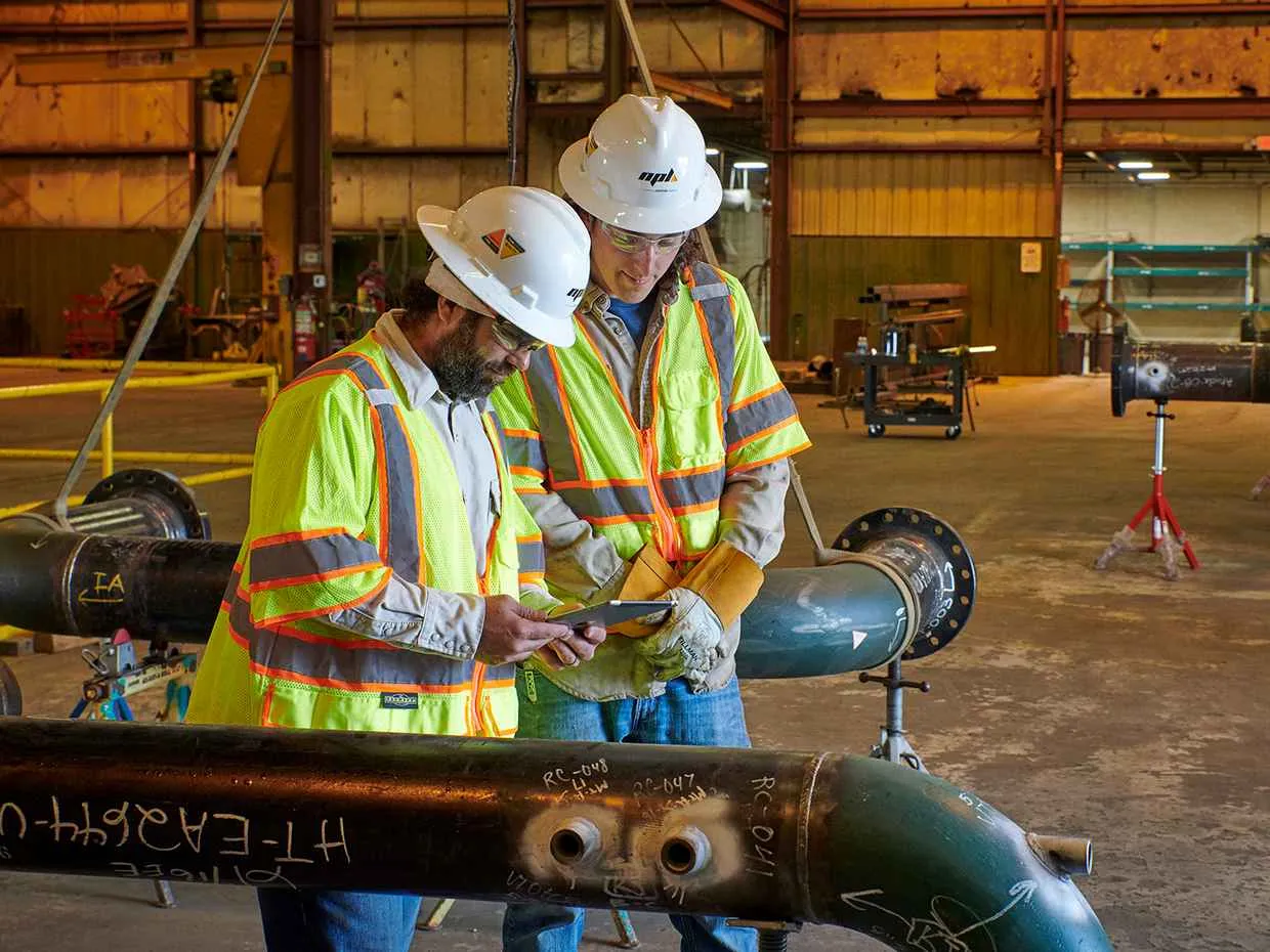 Two pipefitters or construction engineers, wearing hard hats and high-visibility vests, stand in a fabrication shop reviewing information on a digital tablet while inspecting a section of pipe with welded flanges and handwritten markings.