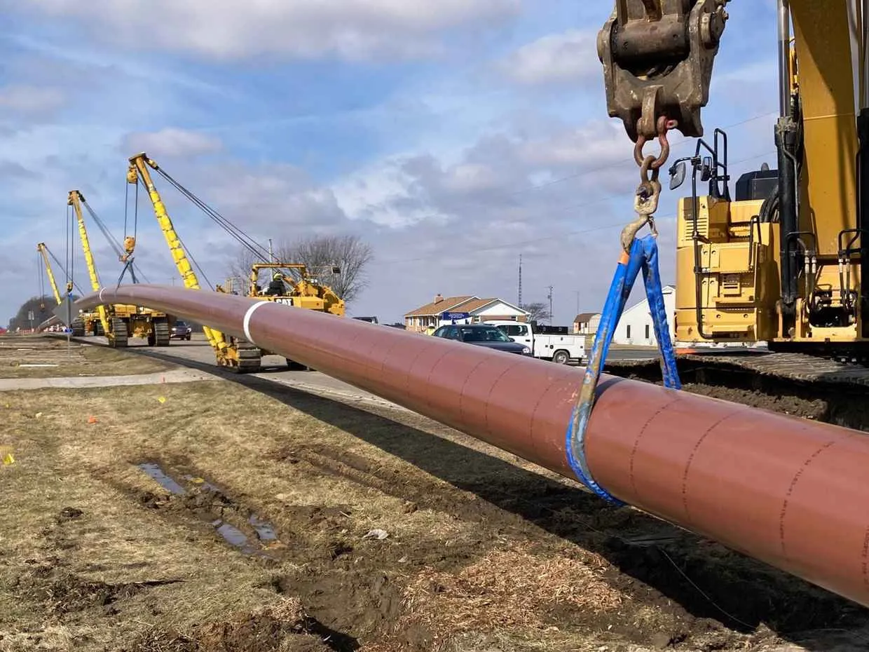 Close-up of a large, rust-colored steel pipe being lowered into a trench using a blue lifting sling attached to a yellow construction crane or pipelayer, with more equipment visible in the background along a suburban road.