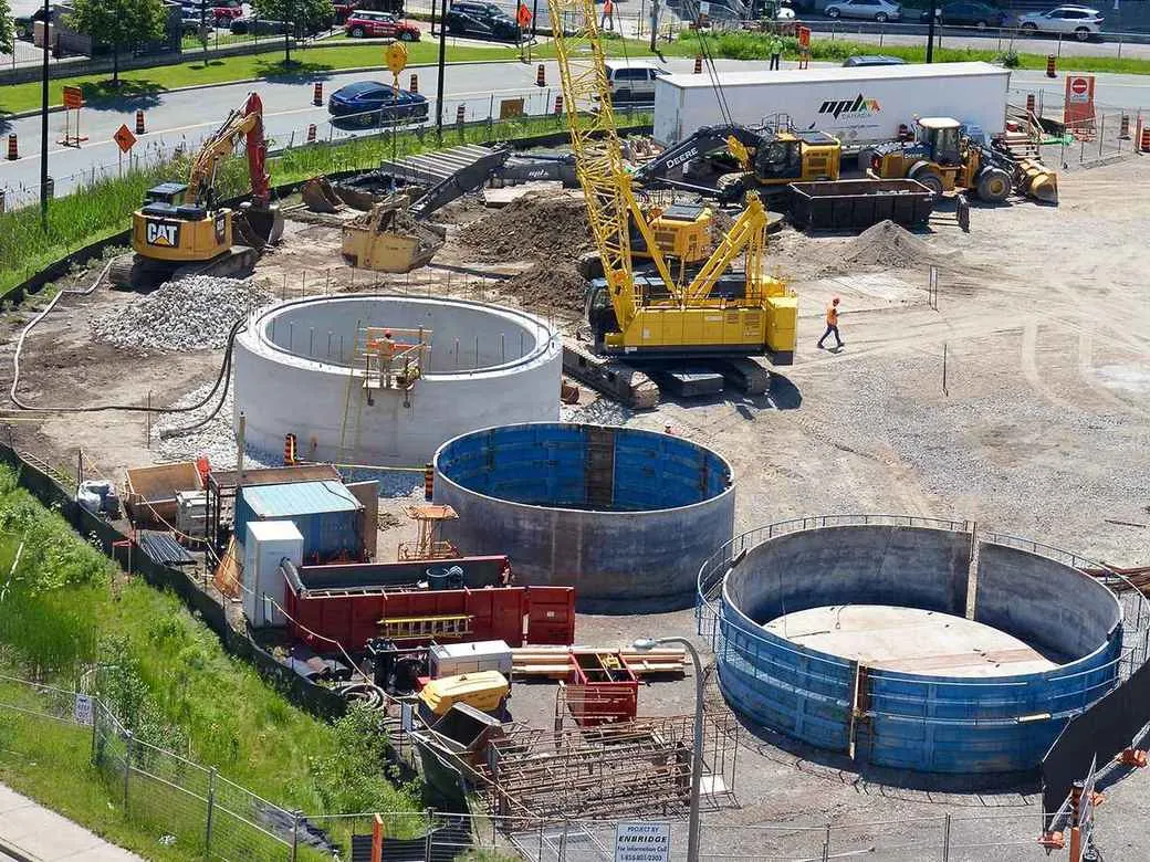 An aerial view of a civil construction site showing large circular concrete structures (likely for a shaft or cistern), with yellow cranes and heavy machinery like a CAT excavator, and workers in high-visibility gear.