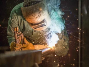 A welder in a darkened welding mask and gloves is working on a metal joint, creating bright sparks and a plume of smoke.