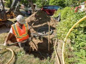 A utility worker in a white hard hat and orange high-visibility vest is standing in a deep trench on a residential property, working to install or repair black cables and yellow flexible pipes.