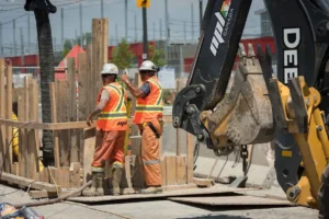 Two construction workers in white hard hats and orange high-visibility vests are standing on a job site next to wooden trench shoring, with the bucket of a large excavator visible on the right.