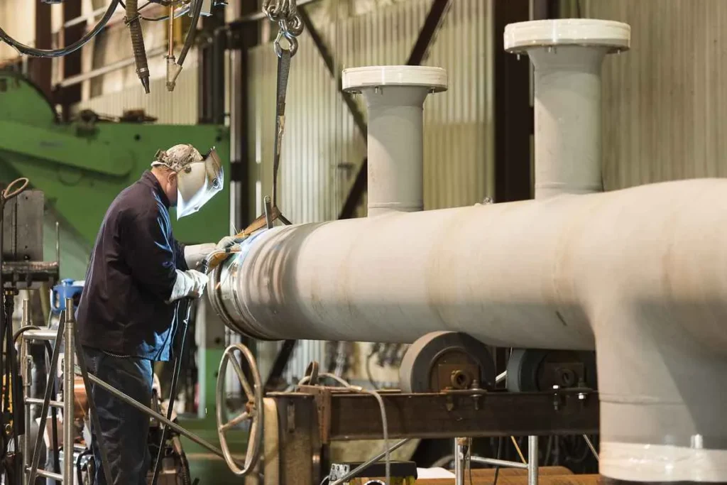 A welder in a darkened welding mask and dark clothing is performing a weld on a large, light grey industrial pipe assembly suspended in a fabrication shop.