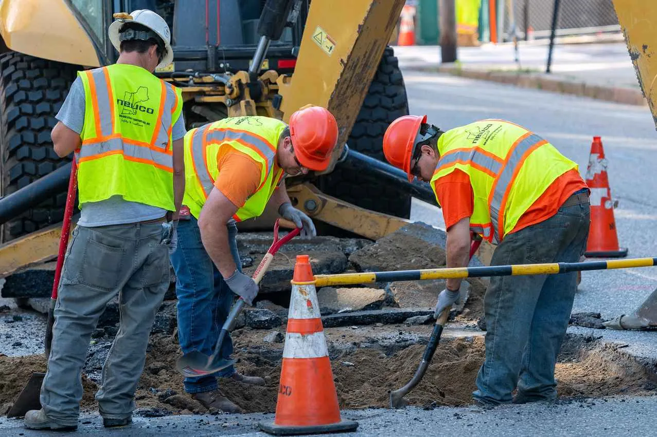 Three construction workers in hard hats and high-visibility vests are using shovels to manually dig a trench in an asphalt road, working around an orange traffic cone and a yellow backhoe parked behind them.