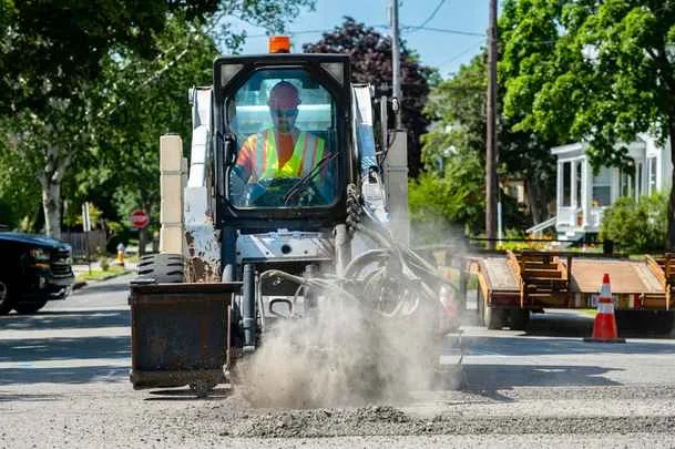 A construction worker in a hard hat and safety vest operates a white skid-steer loader with a bucket attachment, kicking up dust while working on an asphalt street in a residential neighborhood.