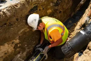A construction worker in a white hard hat and high-visibility vest is bent over in a deep dirt trench, using a clamp or wrench to connect two sections of thick, black utility pipe.