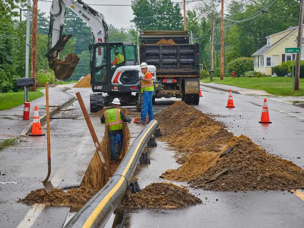 A construction crew is performing utility work on a residential street. A white Bobcat excavator is parked behind a dump truck, while two workers in hard hats and safety vests stand near a long, open trench containing a black and yellow pipe. Piles of dirt and orange traffic cones surround the trench.