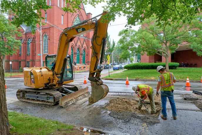 A bright yellow CAT excavator is parked on an asphalt road next to two construction workers who are manually digging in a trench. In the background is a large red brick church and a city street.