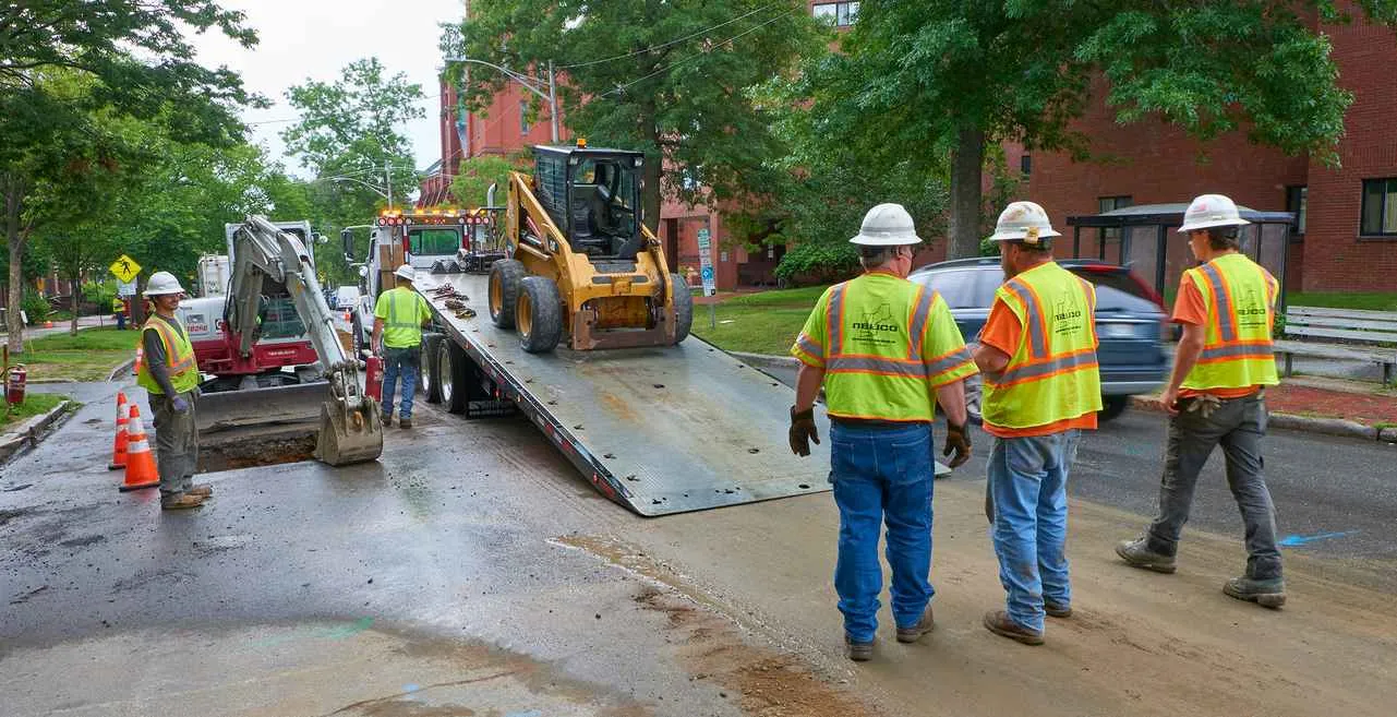 Construction workers in hard hats and high-visibility vests oversee the unloading of a yellow skid-steer loader from a flatbed tow truck parked on an urban street near a utility trench. A small excavator is visible to the left.
